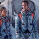 Expressionless astronauts Jo Fowler and Brian Harper stand in front of a crashed space shuttle.